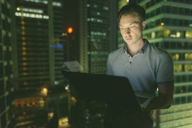 Gece manzaralı cam pencereye dizüstü bilgisayar kullanan yakışıklı bir adamın portresi.