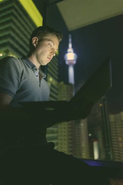 Gece manzaralı cam pencereye dizüstü bilgisayar kullanan yakışıklı bir adamın portresi.