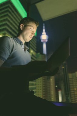 Gece manzaralı cam pencereye dizüstü bilgisayar kullanan yakışıklı bir adamın portresi.