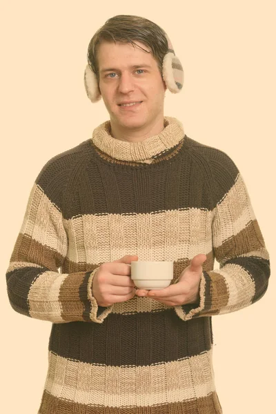 Szczęśliwy przystojny biały mężczyzna nosi zimowe nauszniki i trzyma filiżankę kawy — Zdjęcie stockowe