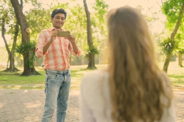 Hombre indio feliz sonriendo y tomando fotos con el teléfono móvil mientras mira a la hermosa mujer enamorada en el tranquilo parque verde — Foto de Stock