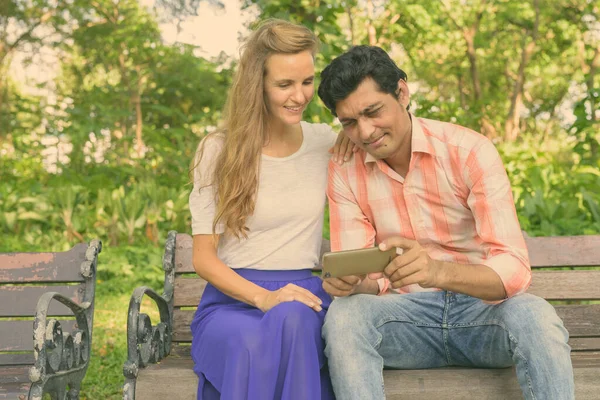 Feliz pareja multi étnica sonriendo mientras se utiliza el teléfono móvil y sentarse juntos en el banco de madera en el amor en el tranquilo parque verde — Foto de Stock