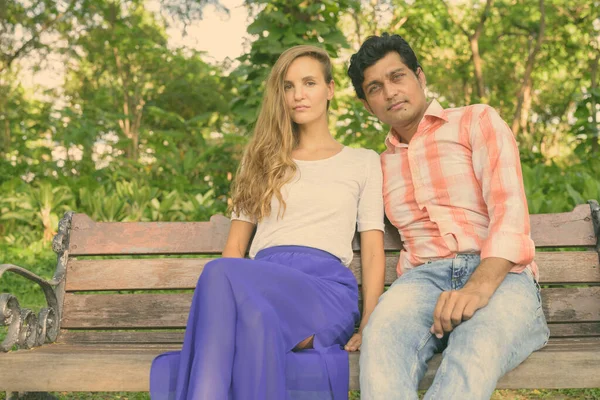 Coppia multietnica seduta su una panchina di legno innamorata in un tranquillo parco verde — Foto Stock