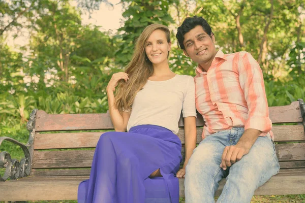 幸せな多民族のカップル笑顔と平和的な緑の公園で愛の木製のベンチに座って — ストック写真