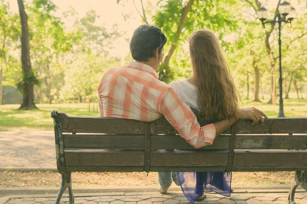 Vista posteriore della coppia multietnica seduta su una panchina di legno innamorata mentre si gode la vista panoramica di un tranquillo parco verde — Foto Stock