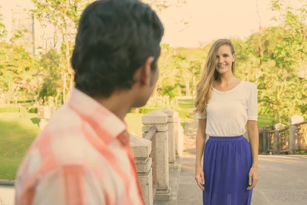Mulher bonita feliz sorrindo enquanto está de pé e olhando para o homem indiano apaixonado na ponte do parque verde pacífico — Fotografia de Stock