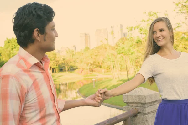 Felice coppia multietnica sorridente mentre si guarda e si tiene per mano insieme sul ponte del tranquillo parco verde — Foto Stock