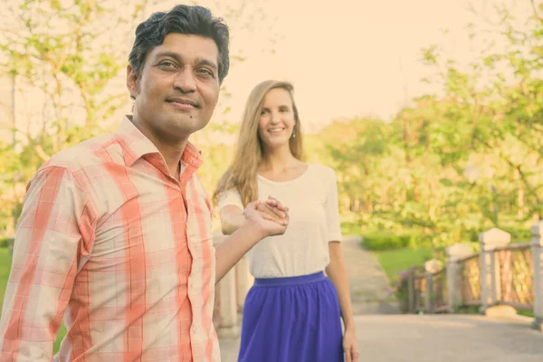 Feliz pareja multiétnica sonriendo con el hombre indio liderando a una hermosa mujer mientras se toman de la mano en el puente del tranquilo parque verde — Foto de Stock