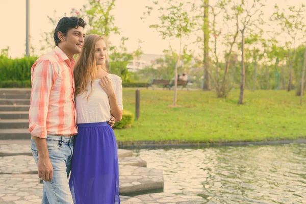 Pareja multiétnica de pie y pensando en el camino de piedra en el medio del lago contra la vista del tranquilo parque verde — Foto de Stock