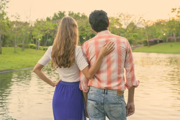 Vista posteriore della coppia multietnica in piedi e godendo la vista panoramica del lago in un tranquillo parco verde insieme — Foto Stock