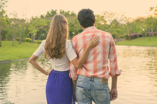 Vista trasera de la pareja multiétnica de pie y disfrutando de la vista panorámica del lago en el tranquilo parque verde juntos — Foto de Stock