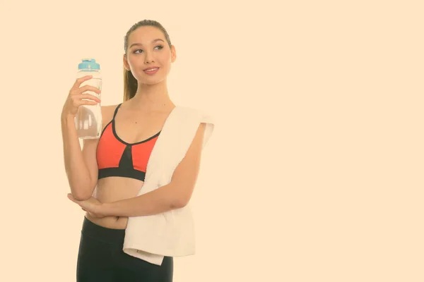 Studio shot van jong gelukkig mooi aziatisch vrouw glimlachen en denken terwijl het houden van water fles klaar voor gym — Stockfoto