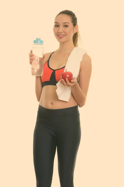 Studio shot młodej szczęśliwej pięknej Azjatki uśmiechającej się i stojącej trzymając butelkę wody i czerwone jabłko gotowe do siłowni — Zdjęcie stockowe