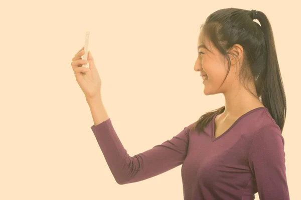 Vista del perfil de la joven adolescente asiática feliz sonriendo mientras toma selfie con teléfono móvil — Foto de Stock