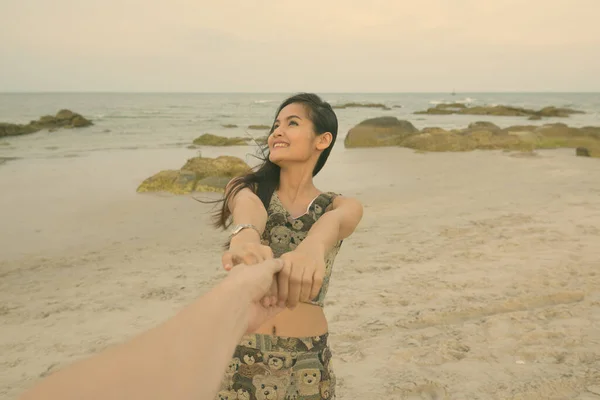 Junge glückliche schöne Asiatin lächelt und denkt, während sie Händchen hält und einen führenden Mann am öffentlichen Strand von Hua Hin in Thailand führt — Stockfoto