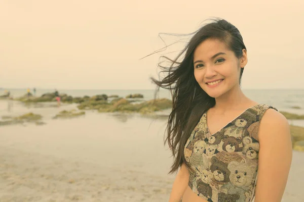 Junge glückliche schöne Asiatin lächelt am windigen öffentlichen Strand von Hua Hin in Thailand — Stockfoto