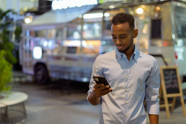 Portrait of handsome black African businessman outdoors in city at night using mobile phone horizontal shot
