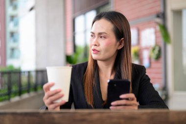 Siyah ceketli, kendine güvenen Taylandlı bir iş kadınının açık hava portresi modern ofis ortak alanında mola veriyor, telefon kullanarak profesyonellik ve liderlik yayıyor.