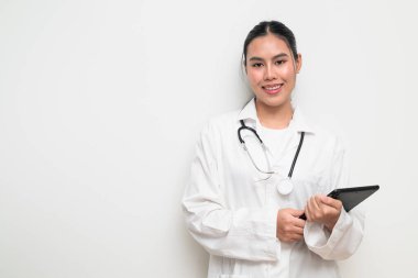 Beyaz üniformalı genç bir Taylandlı kadın doktorun portresi, arka planda steteskop bulunan dijital tablet bir bilgisayar kullanıyor, sağlık reklamcılığı veya tıbbi iletişim için uygun..