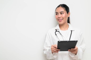 Beyaz üniformalı genç bir Taylandlı kadın doktorun portresi, arka planda steteskop bulunan dijital tablet bir bilgisayar kullanıyor, sağlık reklamcılığı veya tıbbi iletişim için uygun..