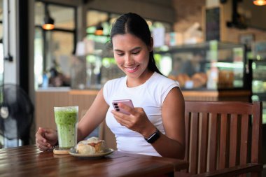 Gülümseyen Asyalı Kadın Kafe 'de Yeşil Buzlu Çay Matcha Latte İçkisi ve Tatlıyla Akıllı Telefon Kullanıyor