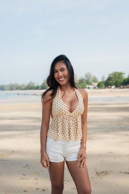 Genç ve güzel Asyalı kadın kumlu sahilde poz veriyor. Açık yaz kıyafetleri giyiyor.