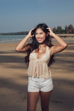 Genç ve güzel Asyalı kadın kumlu sahilde poz veriyor. Açık yaz kıyafetleri giyiyor.