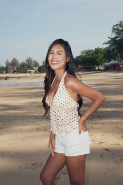 Genç ve güzel Asyalı kadın kumlu sahilde poz veriyor. Açık yaz kıyafetleri giyiyor.