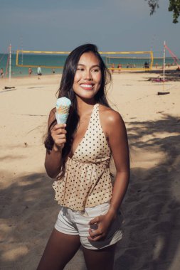 Güneşli bir sahilde dururken elinde mavi dondurma külahıyla gülümseyen genç bir kadının portresi.