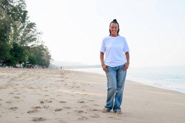 Sıcak gün ışığında kumlu sahilde poz veren kendinden emin yaşlı bir kadın.