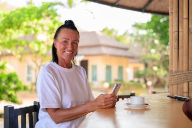 Olgun bir kadın tropikal ortamda kahvenin tadını çıkarırken telefonla mesajlaşıyor..