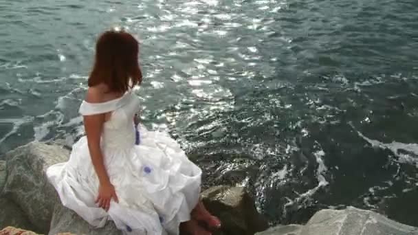 Deniz tarafından kayaya oturan beyaz elbiseli genç ve güzel kadın — Stok video