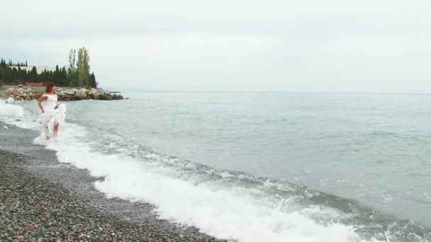 Jonge vrouw In witte jurk loopt langs het strand lopen in zee — Stockvideo
