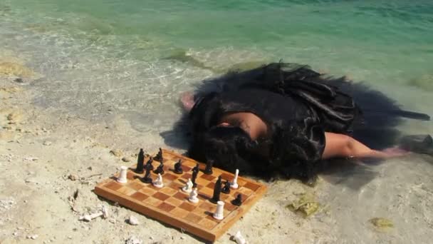 Ung kvinna i svart klänning med styrelsen för schack nära Lake — Stockvideo