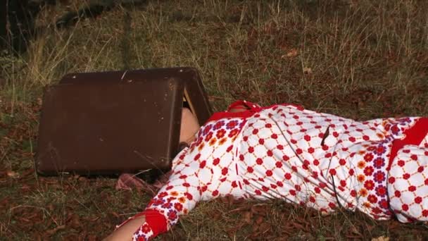 Konstig kvinna i vitprickig klänning liggande på äng med resväska på huvudet i naturen — Stockvideo