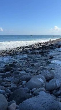 Dalgalar mavi gökyüzünün altındaki kayalık bir kıyı şeridinde yavaşça kopar.