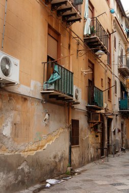 Sunlit alley with peeling plaster, warm-toned walls, and iron balconies. Outdoor air units, laundry flapping, and scattered debris create a lived-in urban scene full of texture and character.
