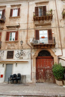 An aged urban building facade featuring rusted balconies, peeling plaster, wooden shutters, and street-level chairs, capturing a rustic European street scene with character and charm.