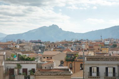 Geniş, güneşli bir şehir manzarası, üzerinde yumuşak bulutlar bulunan engebeli bir dağ zeminine doğru uzanan terracotta çatıları ve pastel binalar içerir.. 
