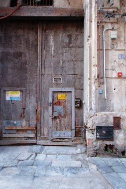 A weathered wooden door and textured stone wall, with exposed pipes, metal panels, and warning signs. The scene evokes aging, history and charm.