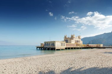 A serene coastal scene features a historic pier and ornate building jutting into calm turquoise waters, with a sandy beach in the foreground and distant mountains under a bright, cloud-dotted sky.