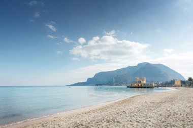 Turkuaz su, yumuşak kumlu kıyılar ve sakin kıyı şeridi boyunca uzanan küçük tarihi bir rıhtım inşaatı tarafından vurgulanan uzak dağlarla sakin bir sahil manzarası..