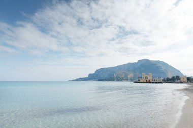 Turkuaz su, yumuşak kumlu kıyılar ve sakin kıyı şeridi boyunca uzanan küçük tarihi bir rıhtım inşaatı tarafından vurgulanan uzak dağlarla sakin bir sahil manzarası..