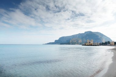 Turkuaz su, yumuşak kumlu kıyılar ve sakin kıyı şeridi boyunca uzanan küçük tarihi bir rıhtım inşaatı tarafından vurgulanan uzak dağlarla sakin bir sahil manzarası..