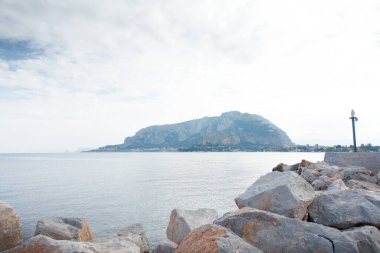 Sakin bir deniz manzarası sakin bir körfez boyunca kayalık bir dalgakıranı, uzak bir adayı ve parlak, parçalı bulutlu bir gökyüzünün altında hafif dalgaları gösterir. Seyahat, doğa ve kıyı yaşam tarzı görselleri için mükemmel.