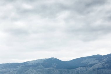 Puslu ve bulutlu bir gökyüzünün altında sakin bir dağ manzarası. Yuvarlanan mavi-gri tepeler doğa, seyahat ve açık hava yaşam tarzı projeleri için uygun sakin ve geniş bir sahne oluşturur..