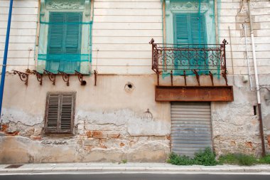 Yıpranmış bir bina cephesinde turkuaz panjurlar, dekoratif demir bir balkon, paslı korkuluklar ve çökmekte olan bir duvar bulunur. Yıpranmış dış görünüş, sakin bir sokak manzarası boyunca kentsel bozulmayı ve tarihi cazibeyi çağrıştırıyor..