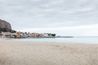 A tranquil seaside town along a sandy beach, with a calm harbor, pastel buildings, boats docked by the piers, and a rocky cliff to the left. Perfect for travel, lifestyle, and vacation scenes.