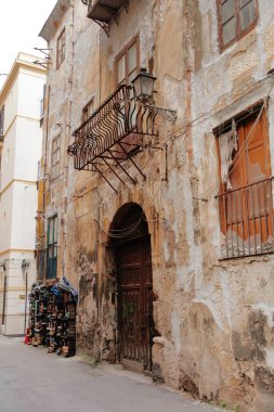 A narrow street in a weathered district shows a crumbling plaster wall, an ornate iron balcony, a wooden door, and a stack of books beside the wall, capturing urban decay and rustic charm.