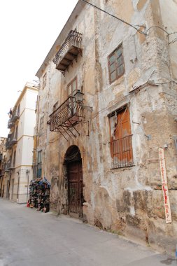 A narrow street in a weathered district shows a crumbling plaster wall, an ornate iron balcony, a wooden door, and a stack of books beside the wall, capturing urban decay and rustic charm.